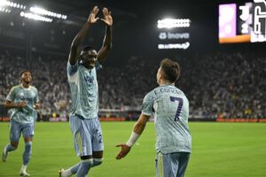 A imagem mostra jogadores de futebol comemorando um gol em um estádio. Um jogador, à esquerda, levanta as mãos em celebração, enquanto outro jogador, à direita, se aproxima para cumprimentá-lo. O fundo é composto por uma multidão de torcedores e as luzes do estádio estão acesas. Os jogadores estão vestindo uniformes azuis claros.