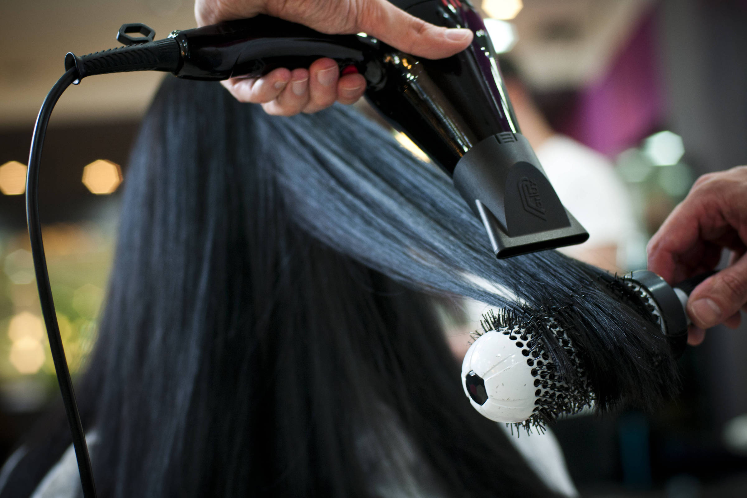 A imagem mostra uma pessoa secando o cabelo com um secador. O cabelo é longo e liso, e está sendo modelado com uma escova redonda. A mão que segura o secador é visível, assim como a escova que está em contato com o cabelo. O fundo é desfocado, com luzes suaves que sugerem um ambiente de salão de beleza.