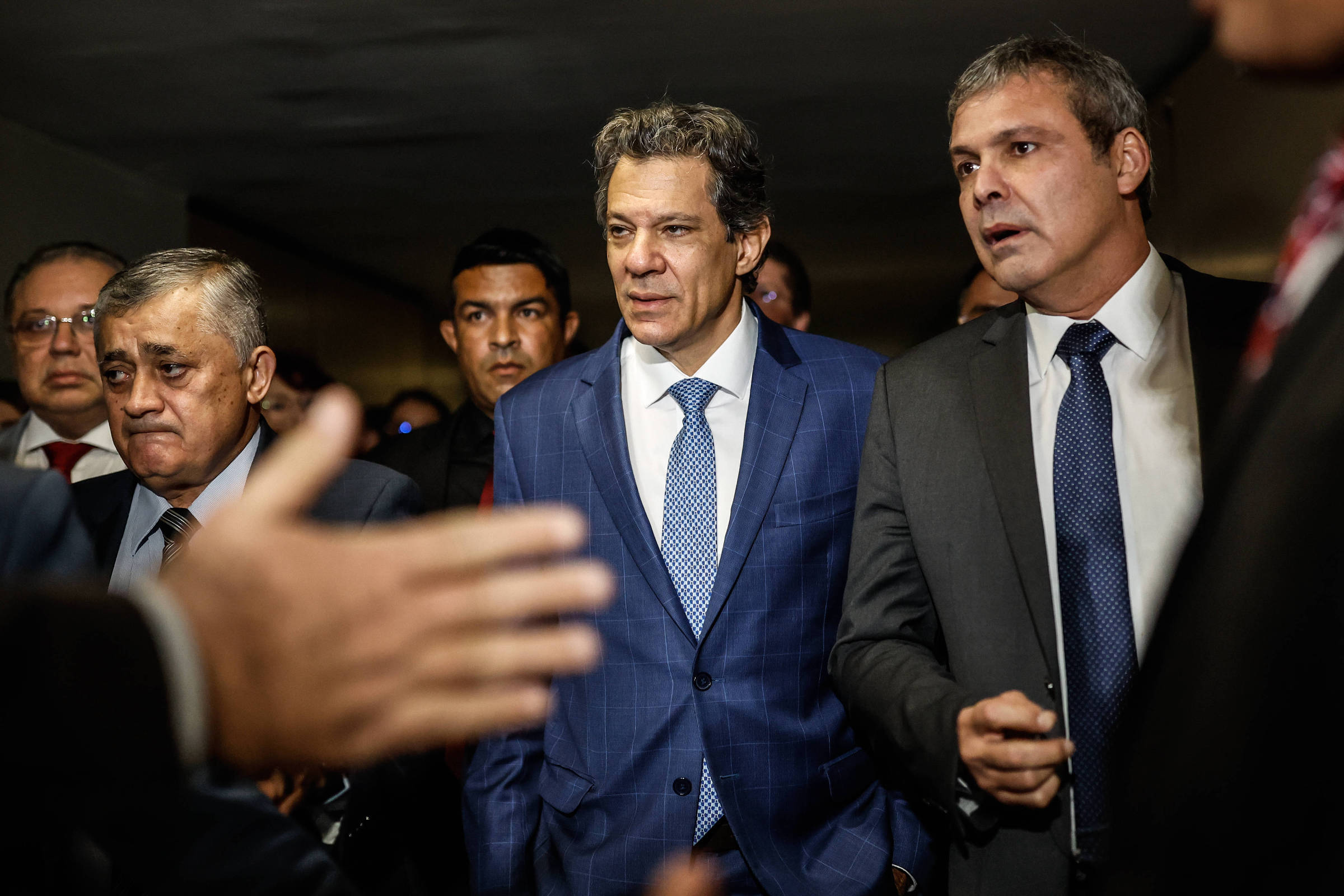 A imagem mostra um grupo de homens em um ambiente formal. No centro, um homem com cabelo ondulado e terno azul claro parece estar em uma conversa séria. À sua esquerda, um homem de terno escuro e gravata clara também está envolvido na conversa. Outros homens ao fundo observam a cena, alguns com expressões sérias. O ambiente parece ser um corredor ou sala de reuniões.