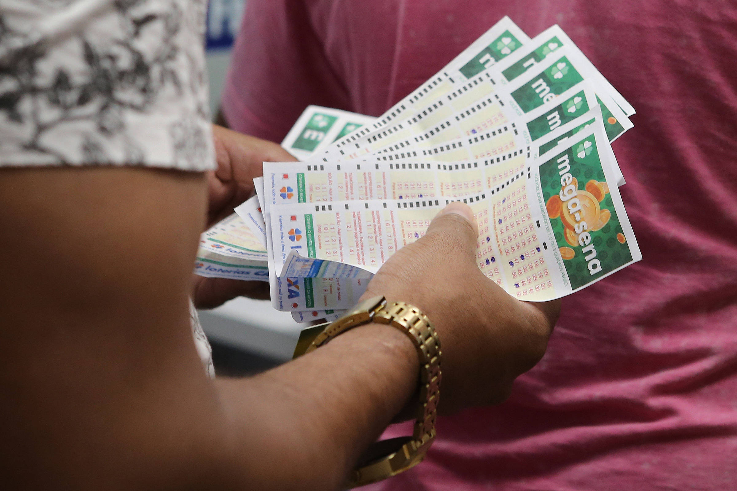 A imagem mostra uma pessoa segurando vários bilhetes de loteria. Os bilhetes são coloridos, com informações visíveis sobre números e jogos. Ao fundo, é possível ver outra pessoa com uma camiseta rosa. A mão que segura os bilhetes é adornada com uma pulseira dourada.