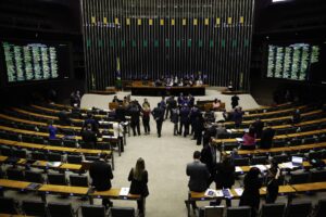 A imagem mostra o plenário da Câmara dos Deputados do Brasil, com várias pessoas em pé e sentadas. Ao fundo, há uma mesa com representantes e bandeiras do Brasil. As paredes laterais estão equipadas com painéis eletrônicos que exibem informações. O ambiente é amplo, com cadeiras dispostas em fileiras e um piso claro.