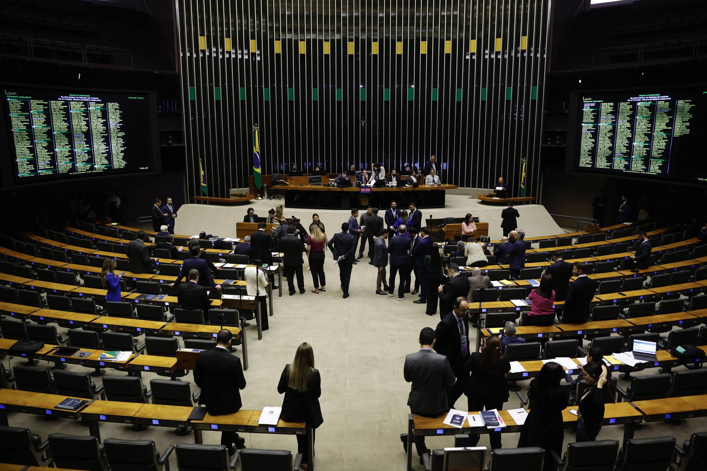 A imagem mostra o plenário da Câmara dos Deputados do Brasil, com várias pessoas em pé e sentadas. Ao fundo, há uma mesa com representantes e bandeiras do Brasil. As paredes laterais estão equipadas com painéis eletrônicos que exibem informações. O ambiente é amplo, com cadeiras dispostas em fileiras e um piso claro.