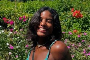 Uma mulher com cabelo solto e escuro sorri em um jardim cheio de flores coloridas. Ela usa uma blusa de alças e um colar. O fundo é composto por várias plantas e flores, predominantemente rosas e outras cores vibrantes, criando um ambiente alegre e natural.