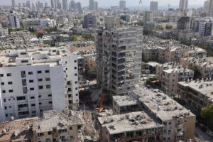 A imagem mostra uma área urbana devastada, com prédios danificados e em ruínas. Um edifício em destaque está parcialmente destruído, com escombros visíveis ao seu redor. Ao fundo, há uma vista da cidade com vários arranha-céus e construções. O céu está claro e a cena parece ser de um local que sofreu um desastre, possivelmente um ataque ou um terremoto.