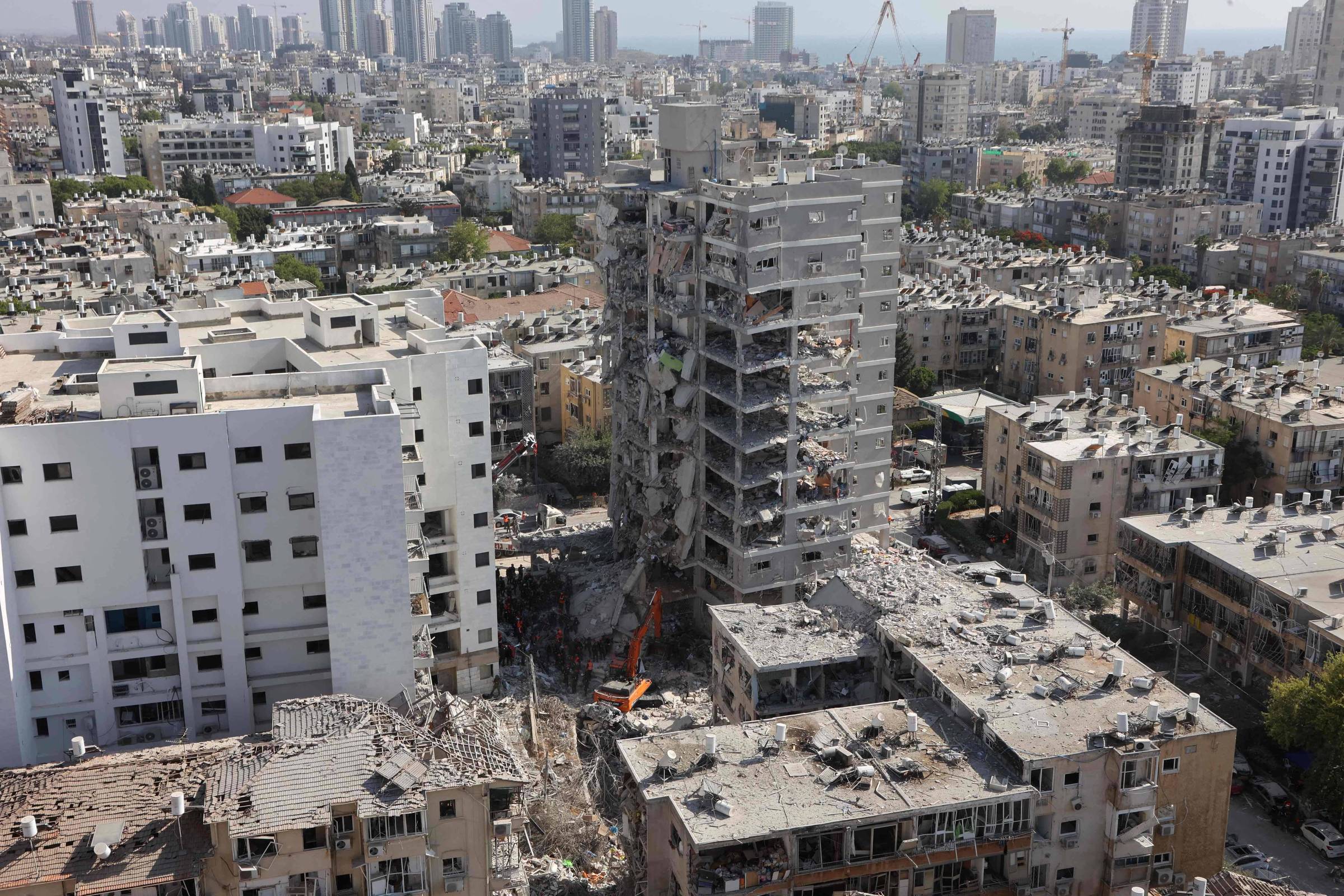 A imagem mostra uma área urbana devastada, com prédios danificados e em ruínas. Um edifício em destaque está parcialmente destruído, com escombros visíveis ao seu redor. Ao fundo, há uma vista da cidade com vários arranha-céus e construções. O céu está claro e a cena parece ser de um local que sofreu um desastre, possivelmente um ataque ou um terremoto.