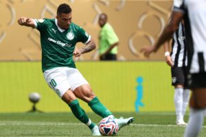 Um jogador de futebol está em campo, vestindo uma camisa verde com detalhes brancos e o número 10. Ele está prestes a chutar a bola, que é colorida. Ao fundo, há um jogador adversário e uma pessoa em uma camisa verde clara. O campo é bem iluminado e há uma parede com um design simples ao fundo.