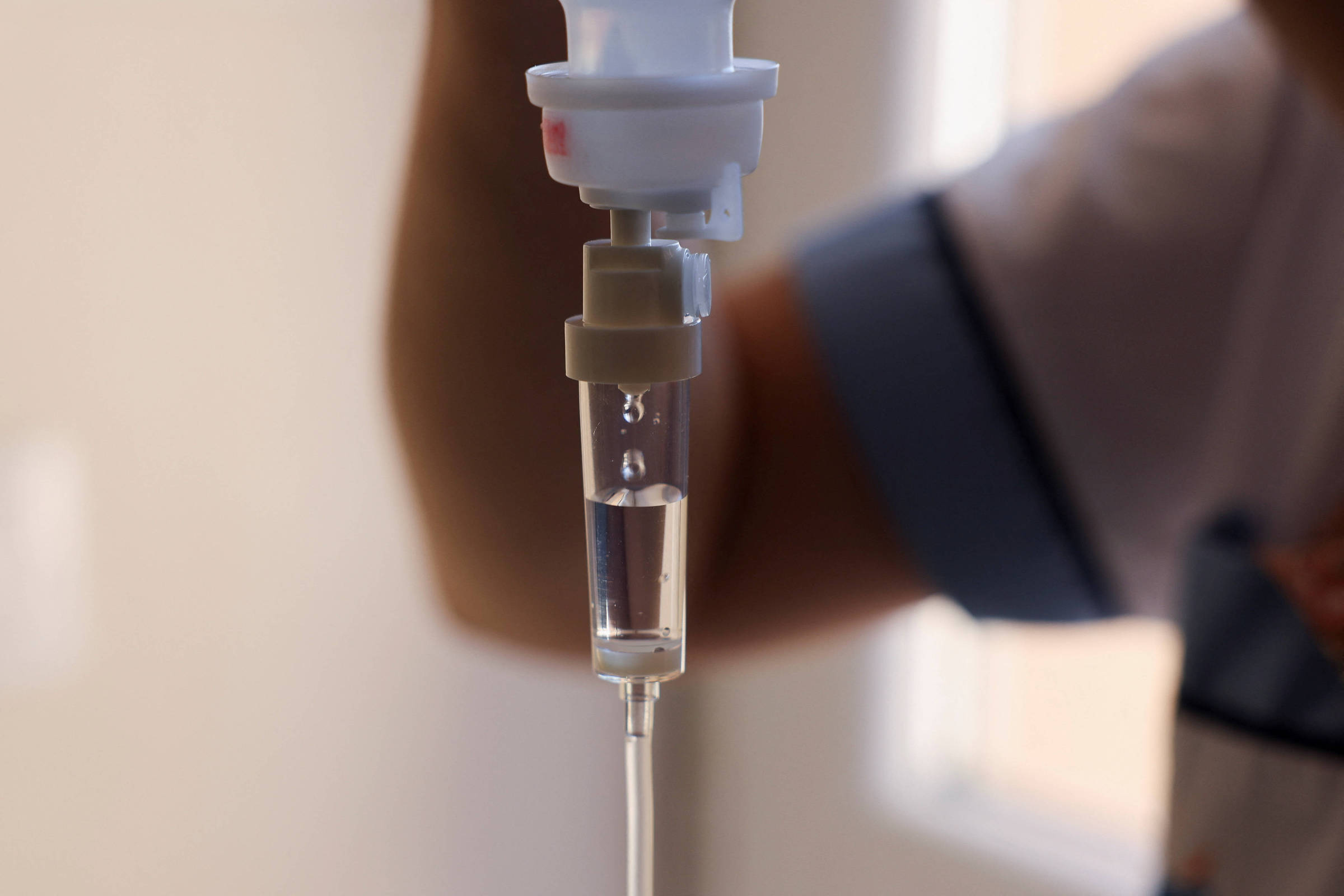 A imagem mostra um frasco de soro intravenoso sendo administrado. O foco está na parte do equipamento onde o líquido está passando, com gotas visíveis descendo pelo tubo. Ao fundo, é possível ver uma mão segurando o equipamento, e a iluminação é suave, com um fundo desfocado.