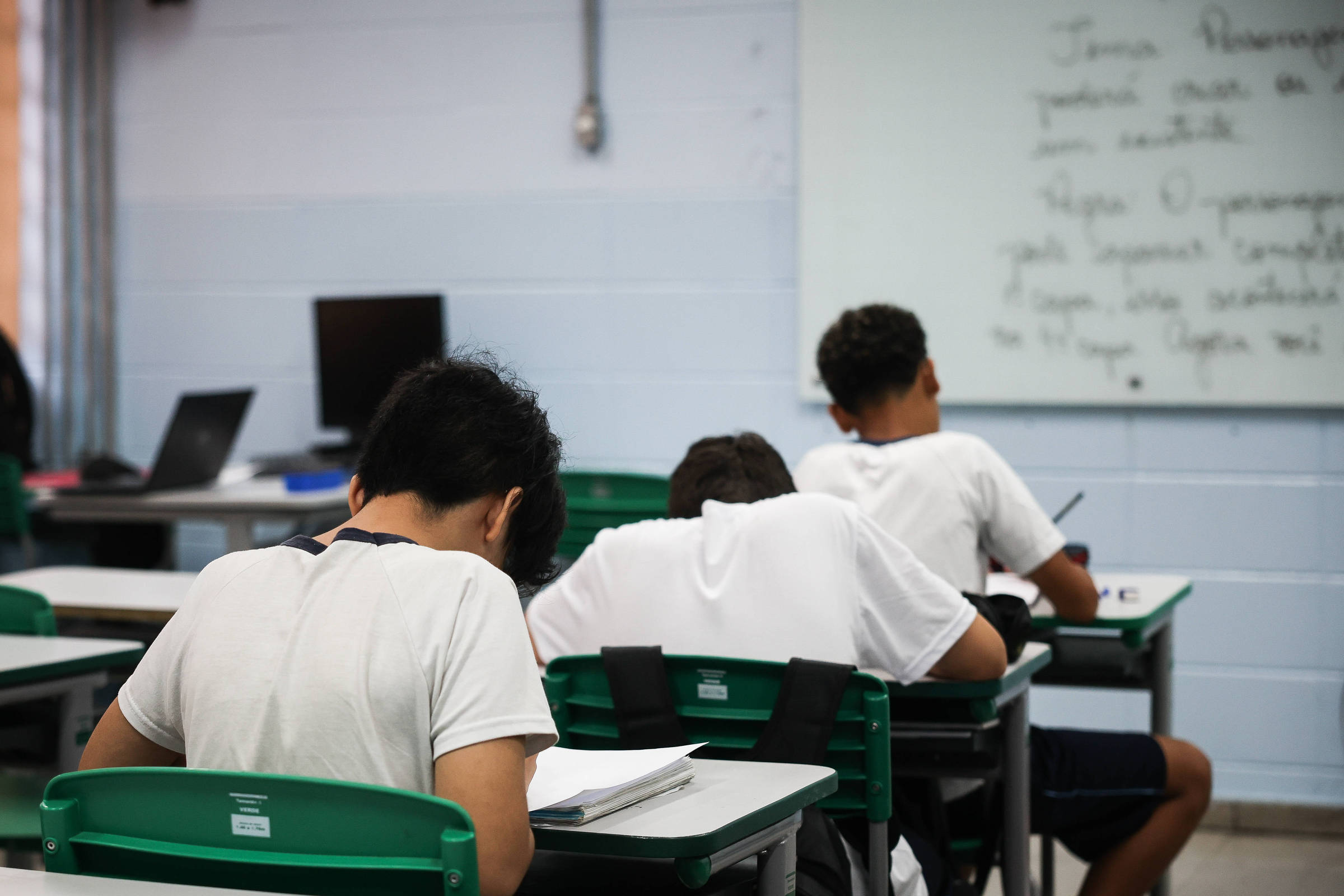 A imagem mostra três estudantes sentados em mesas em uma sala de aula. Eles estão com a cabeça baixa, aparentemente escrevendo ou estudando. Ao fundo, há uma lousa com texto escrito e um computador em uma mesa. As mesas são verdes e os estudantes vestem camisetas brancas.