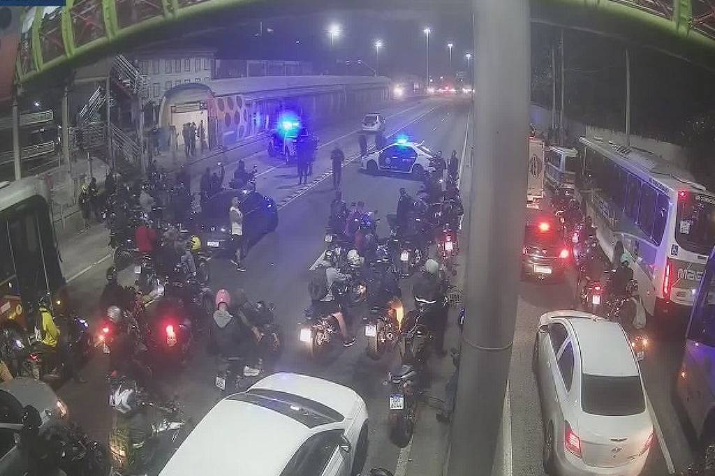 A imagem mostra um congestionamento noturno em uma via, onde há um grande número de motocicletas paradas. Algumas motos estão em destaque, com luzes traseiras acesas. Há também veículos de quatro rodas, incluindo um carro branco e um ônibus. Ao fundo, é possível ver a presença de viaturas policiais com luzes azuis piscando. O ambiente é urbano, com iluminação artificial e uma estrutura elevada visível na parte superior da imagem.