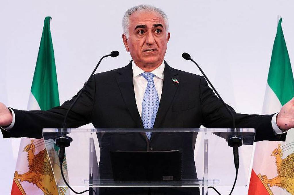 Um homem em um terno escuro está em pé atrás de um púlpito de vidro, gesticulando com as mãos. Ele tem cabelo grisalho e está usando uma gravata clara. Ao fundo, há bandeiras do Irã, com cores verde, branco e vermelho, dispostas em ambos os lados do homem. O ambiente parece ser uma conferência ou evento formal.