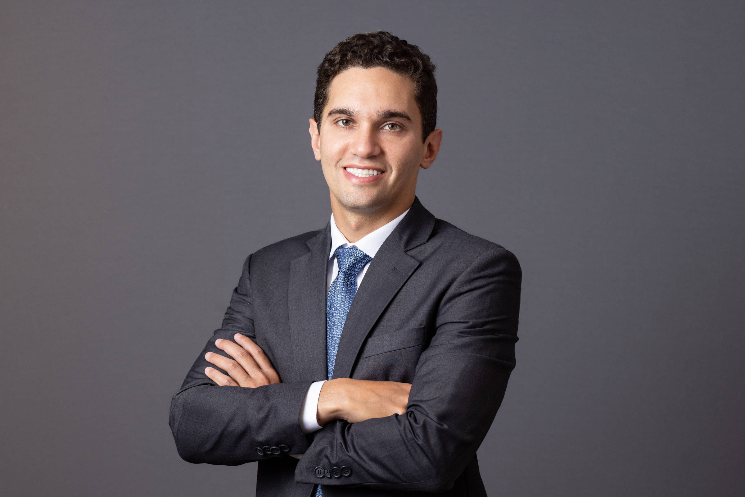 Um homem está posando para a foto, com um sorriso no rosto. Ele está vestindo um terno escuro e uma camisa branca, com uma gravata azul. O fundo da imagem é cinza, e o homem está com os braços cruzados na frente do corpo.
