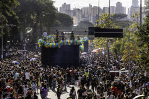 A imagem mostra uma grande multidão reunida em um evento ao ar livre, com pessoas de várias idades e estilos. No centro, há um caminhão de som decorado com balões coloridos, onde estão duas pessoas em cima, possivelmente se apresentando. Ao fundo, é possível ver uma cidade com prédios altos e árvores ao redor. O céu está claro e ensolarado, e algumas pessoas estão usando guarda-sóis.