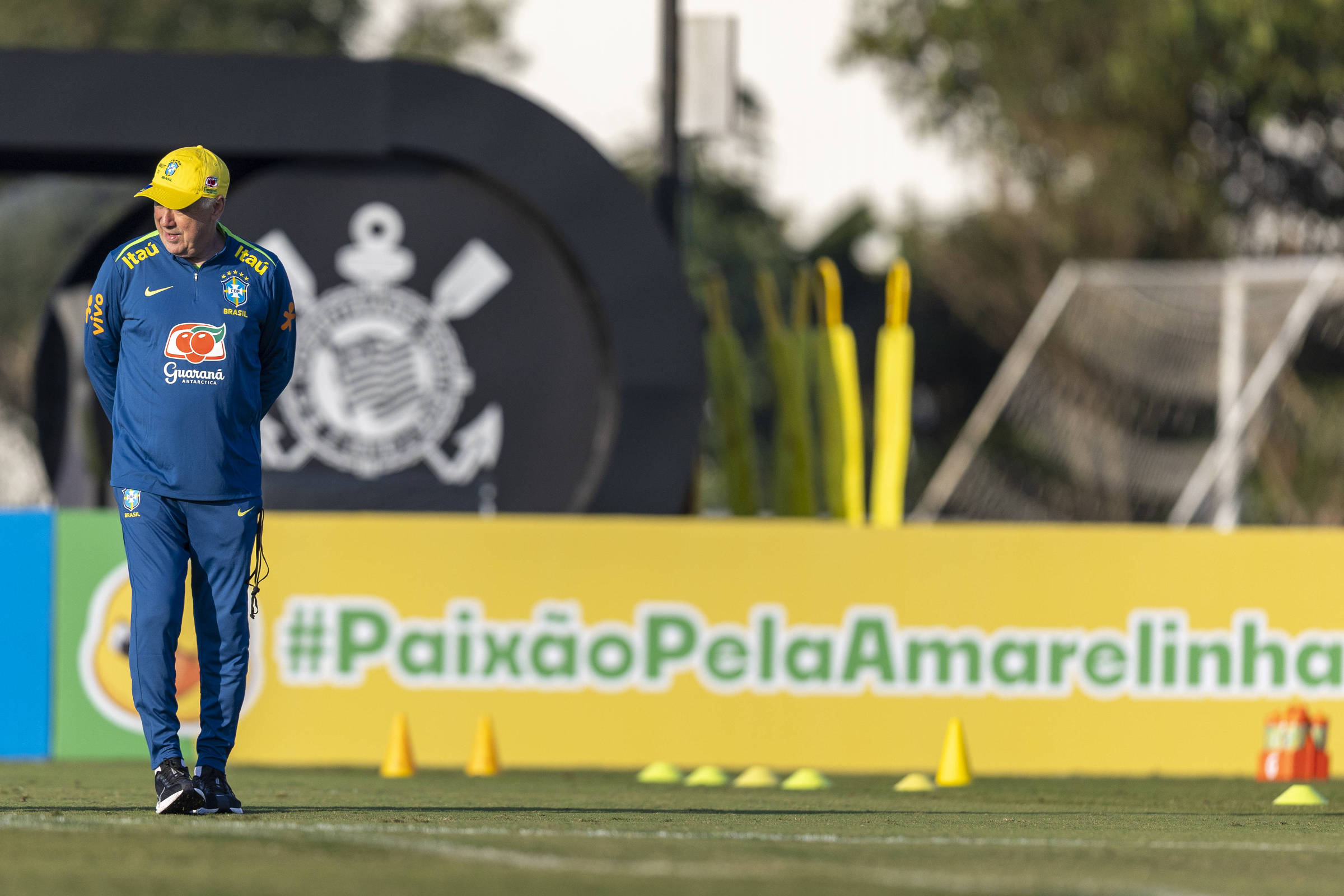 A imagem mostra Carlo Ancelotti, treinador da Seleção Brasileira de futebol, em um campo de treinamento. Ele está vestido com um agasalho azul e uma boné amarelo, observando os cones de treinamento dispostos no gramado. Ao fundo, há uma placa com a frase