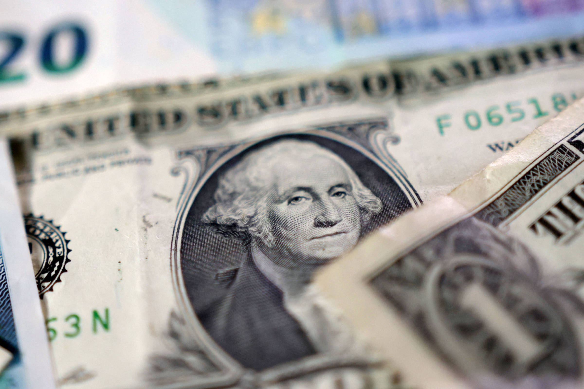 A imagem mostra várias notas de dólar americano em primeiro plano, com destaque para uma nota de um dólar que apresenta o retrato de George Washington. Ao fundo, é possível ver uma nota de vinte dólares parcialmente visível. As notas estão sobrepostas, criando um efeito de desfoque em algumas partes.