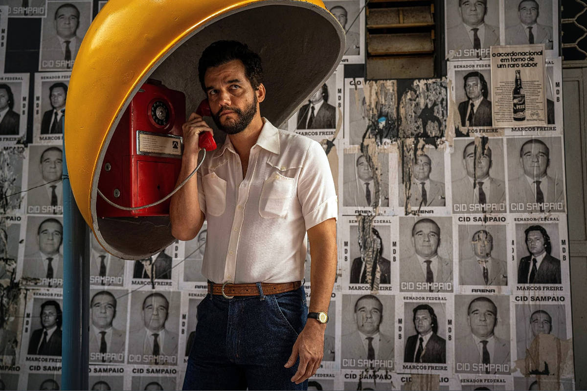 Um homem está em pé, segurando um telefone vermelho em uma cabine telefônica amarela. Ele usa uma camisa branca e jeans escuros, com uma expressão pensativa. Ao fundo, há uma parede coberta com várias fotos em preto e branco de pessoas, algumas com nomes visíveis, como