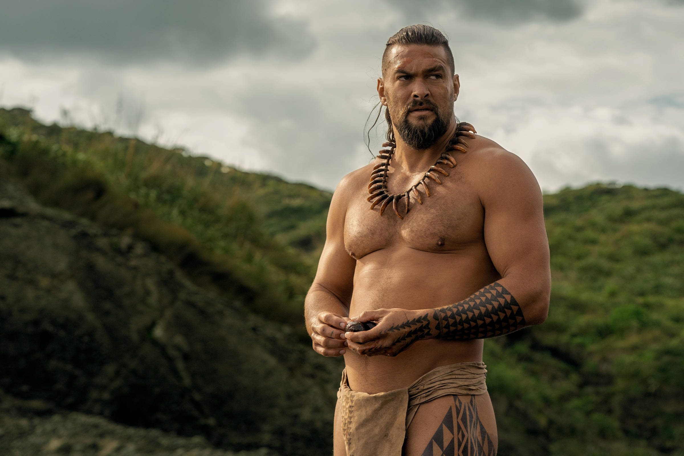Um homem musculoso está em pé em um ambiente natural, com colinas verdes ao fundo. Ele está sem camisa, usando um colar de conchas e uma saia feita de tecido. Seu braço esquerdo possui uma tatuagem tribal. O céu está nublado, sugerindo um clima sombrio.