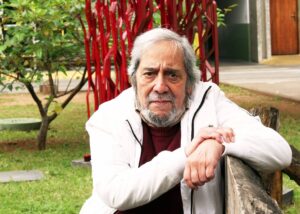 Um homem idoso com cabelo grisalho e barba está sentado em um banco de madeira. Ele usa um suéter branco sobre uma camisa escura e tem uma expressão pensativa. Ao fundo, há um jardim com árvores e uma estrutura vermelha que parece ser uma escultura. O ambiente é bem iluminado e parece ser um espaço ao ar livre.