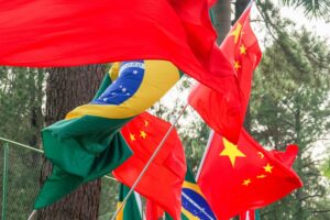 A imagem mostra um grupo de pessoas segurando bandeiras do Brasil e da China em um evento ao ar livre. As pessoas estão alinhadas em uma área verde, com árvores ao fundo. Elas seguram bandeiras vermelhas e verdes, além de faixas com os símbolos dos dois países. O ambiente parece festivo, com várias pessoas sorrindo e interagindo.