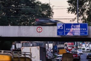 A imagem mostra uma via urbana com tráfego intenso. No primeiro plano, há caminhões e carros em movimento. Acima, uma ponte é visível, com um sinal de altura máxima de 4,8 metros e uma placa indicando