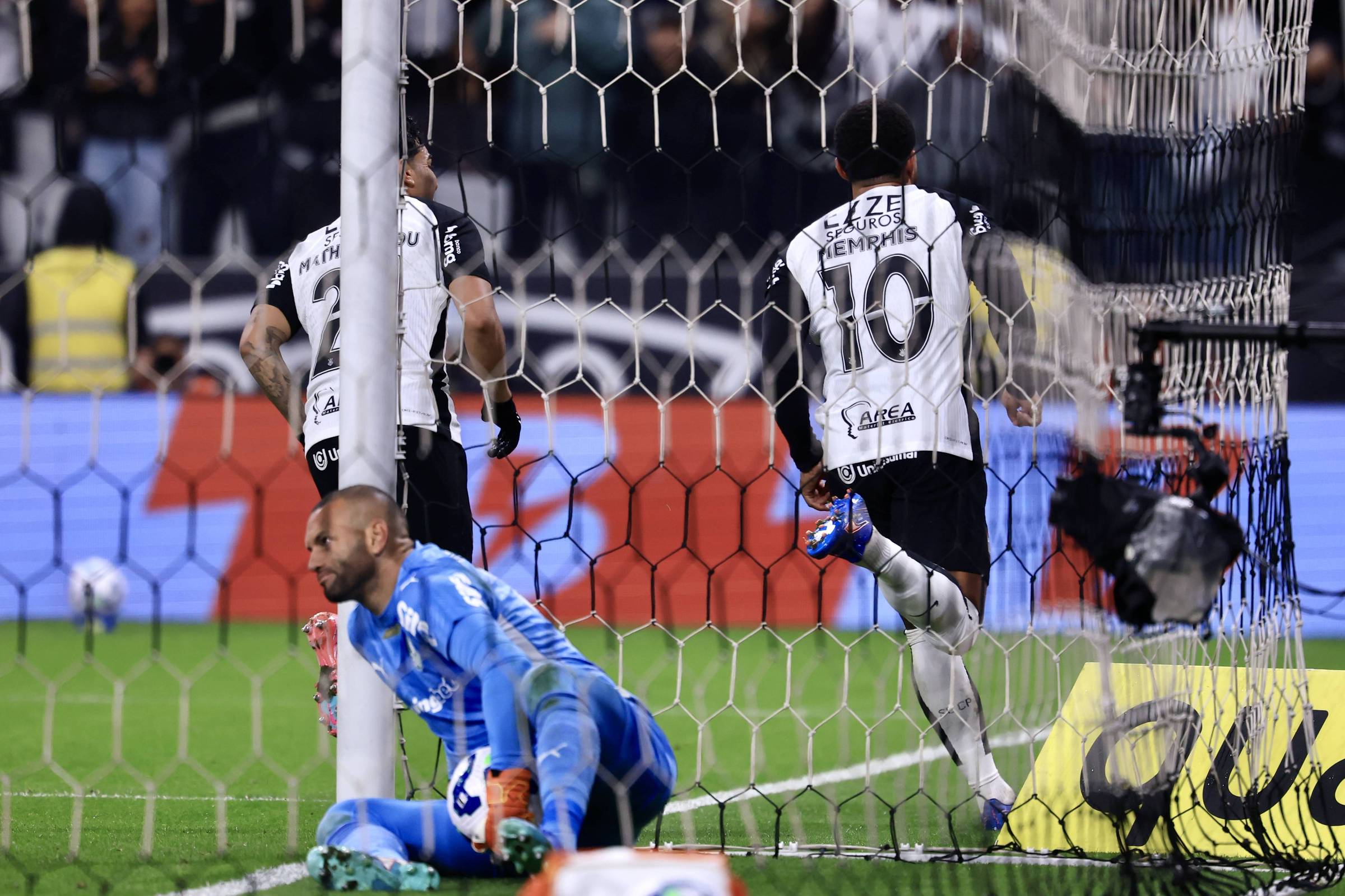 A imagem mostra um momento de um jogo de futebol, onde um jogador, identificado pelo número 10, comemora um gol. Ele está correndo em direção à rede, enquanto dois outros jogadores estão próximos. O goleiro, que está caído no chão, parece estar desapontado. O fundo mostra uma torcida e um campo de futebol iluminado.