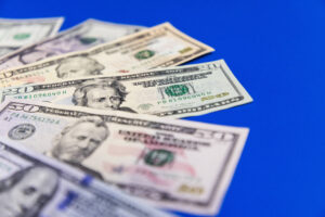 A imagem mostra várias notas de dólar dispostas em uma superfície azul. As notas incluem valores de 20 e 100 dólares, com os rostos de figuras históricas visíveis. As notas estão ligeiramente sobrepostas, criando uma composição que destaca a variedade de denominações.