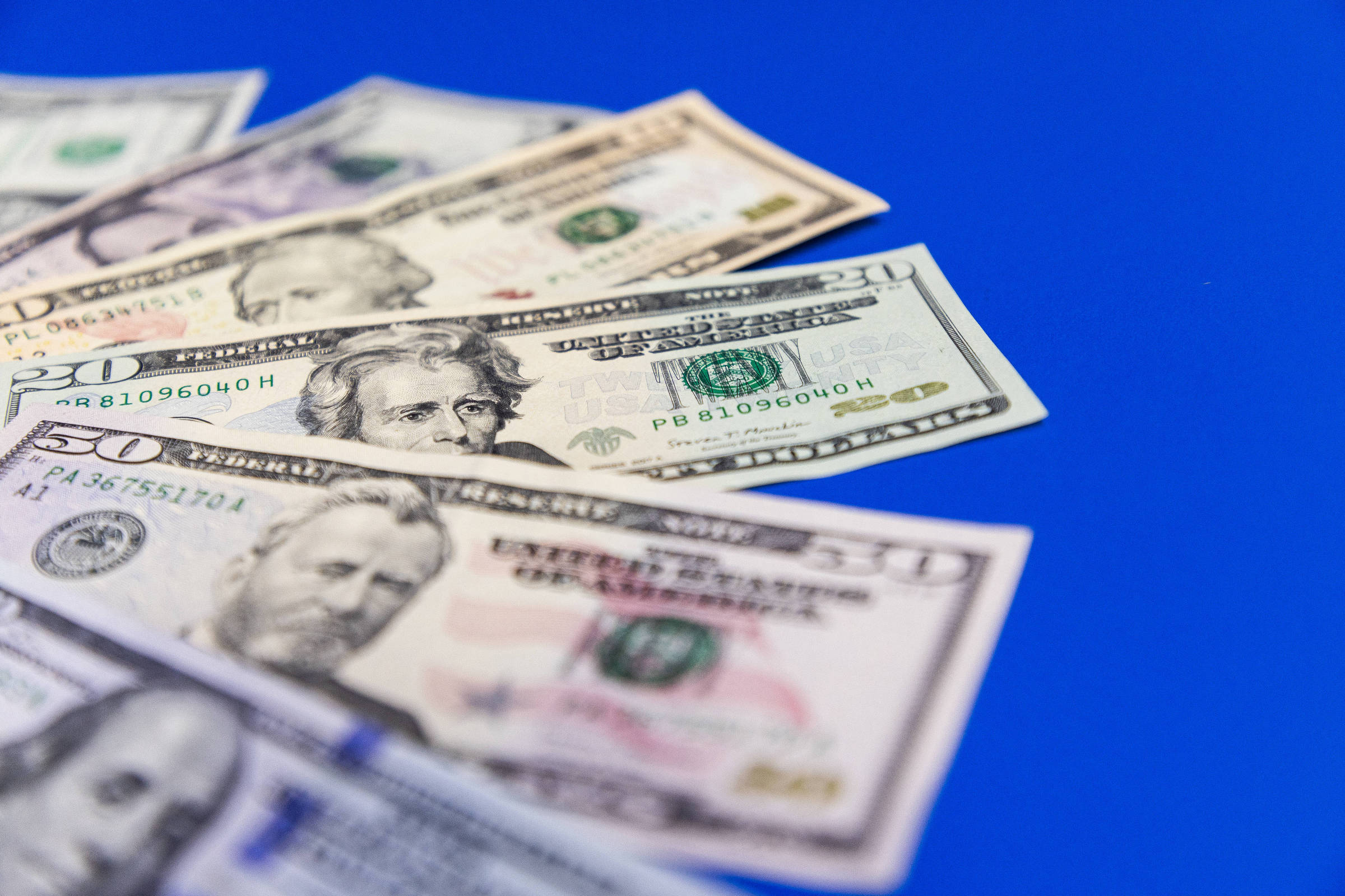 A imagem mostra várias notas de dólar dispostas em uma superfície azul. As notas incluem valores de 20 e 100 dólares, com os rostos de figuras históricas visíveis. As notas estão ligeiramente sobrepostas, criando uma composição que destaca a variedade de denominações.