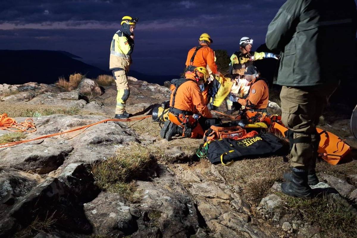 A imagem mostra uma equipe de resgate em uma área montanhosa durante a noite. Vários membros da equipe estão vestidos com uniformes de resgate, incluindo capacetes e coletes refletivos. Eles estão reunidos em torno de uma pessoa que parece estar recebendo atendimento. Há cordas e equipamentos de resgate espalhados pelo chão, e a iluminação é baixa, com nuvens escuras ao fundo.