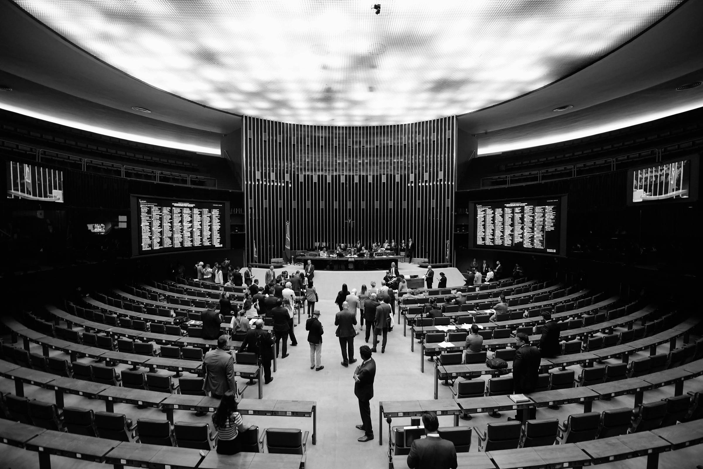 Imagem do plenário da Câmara dos Deputados do Brasil, com várias pessoas em pé e sentadas, algumas interagindo. O ambiente é amplo, com cadeiras dispostas em fileiras e uma mesa central onde estão os deputados. As paredes são decoradas com painéis e há telas digitais exibindo informações. O teto é iluminado com uma luz suave.