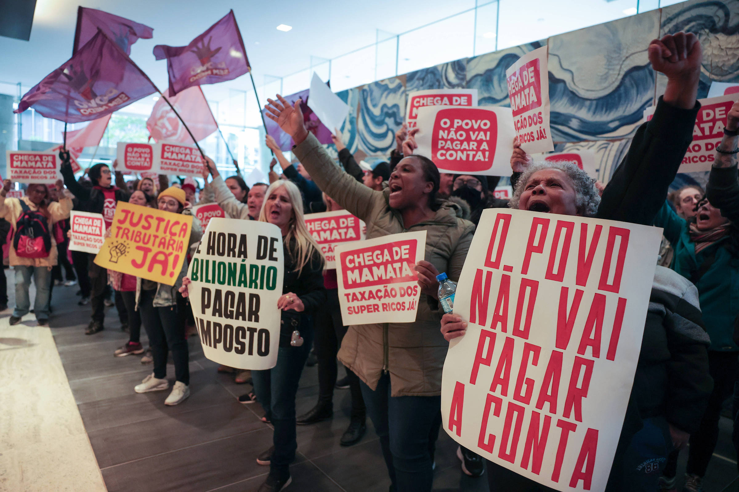 A imagem mostra um grupo de pessoas em um protesto, segurando cartazes e bandeiras. Os cartazes contêm mensagens como