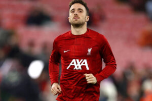 Um jogador de futebol está correndo em um campo, vestindo um uniforme vermelho do Liverpool. O uniforme tem o logotipo do time e a marca