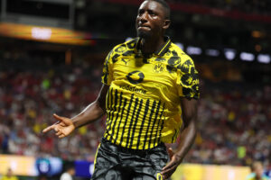 Um jogador de futebol está comemorando um gol. Ele veste uma camisa amarela com detalhes pretos, que é a camisa do Borussia Dortmund. O fundo mostra uma multidão em um estádio, com torcedores visíveis. O jogador parece estar expressando alegria e emoção após a jogada.