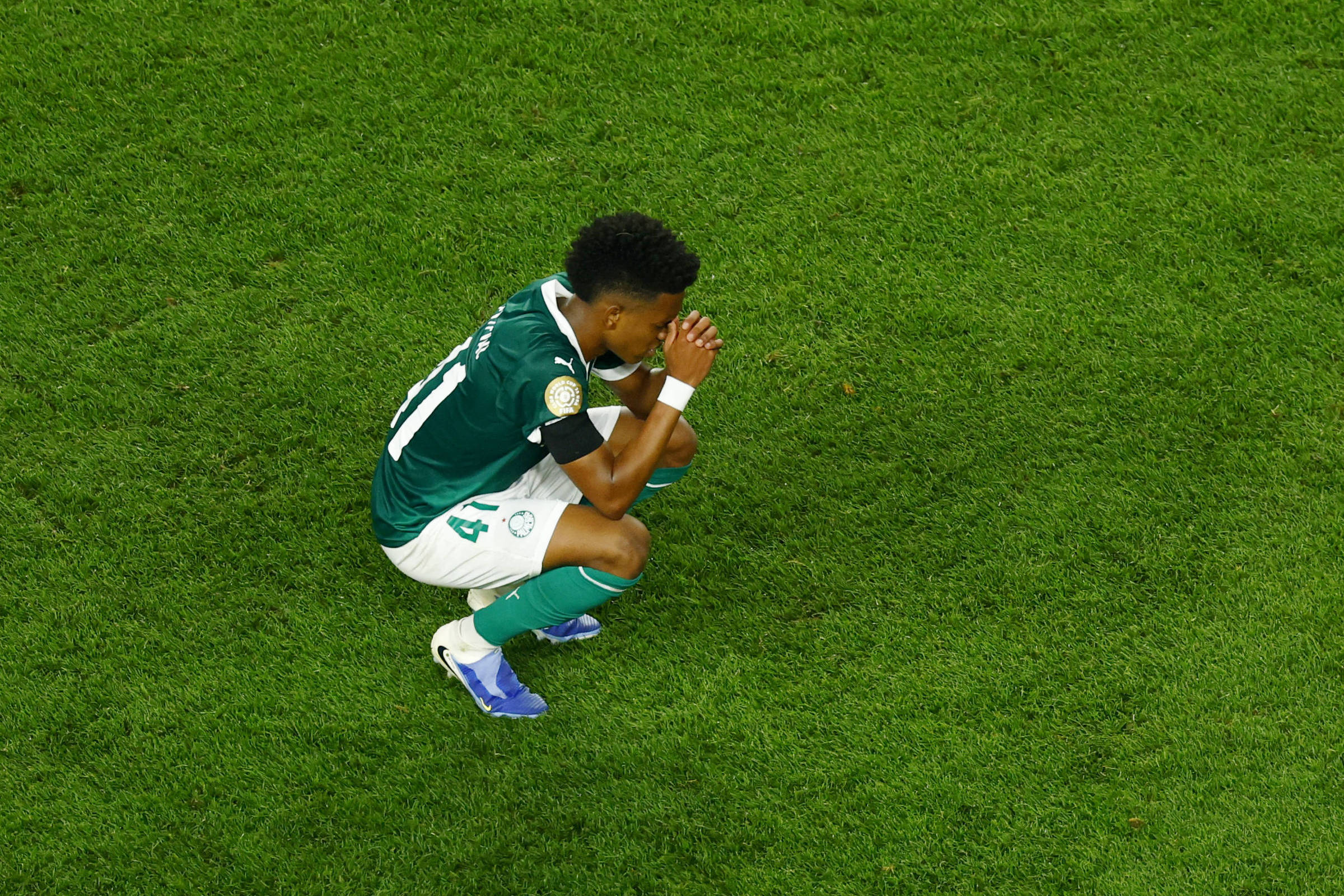 Um jogador de futebol está agachado no campo, com as mãos cobrindo o rosto, em uma expressão de desânimo ou reflexão. Ele veste um uniforme verde e branco, com o número 14 nas costas. O campo é coberto por grama verde, e a imagem captura um momento emocional do atleta.