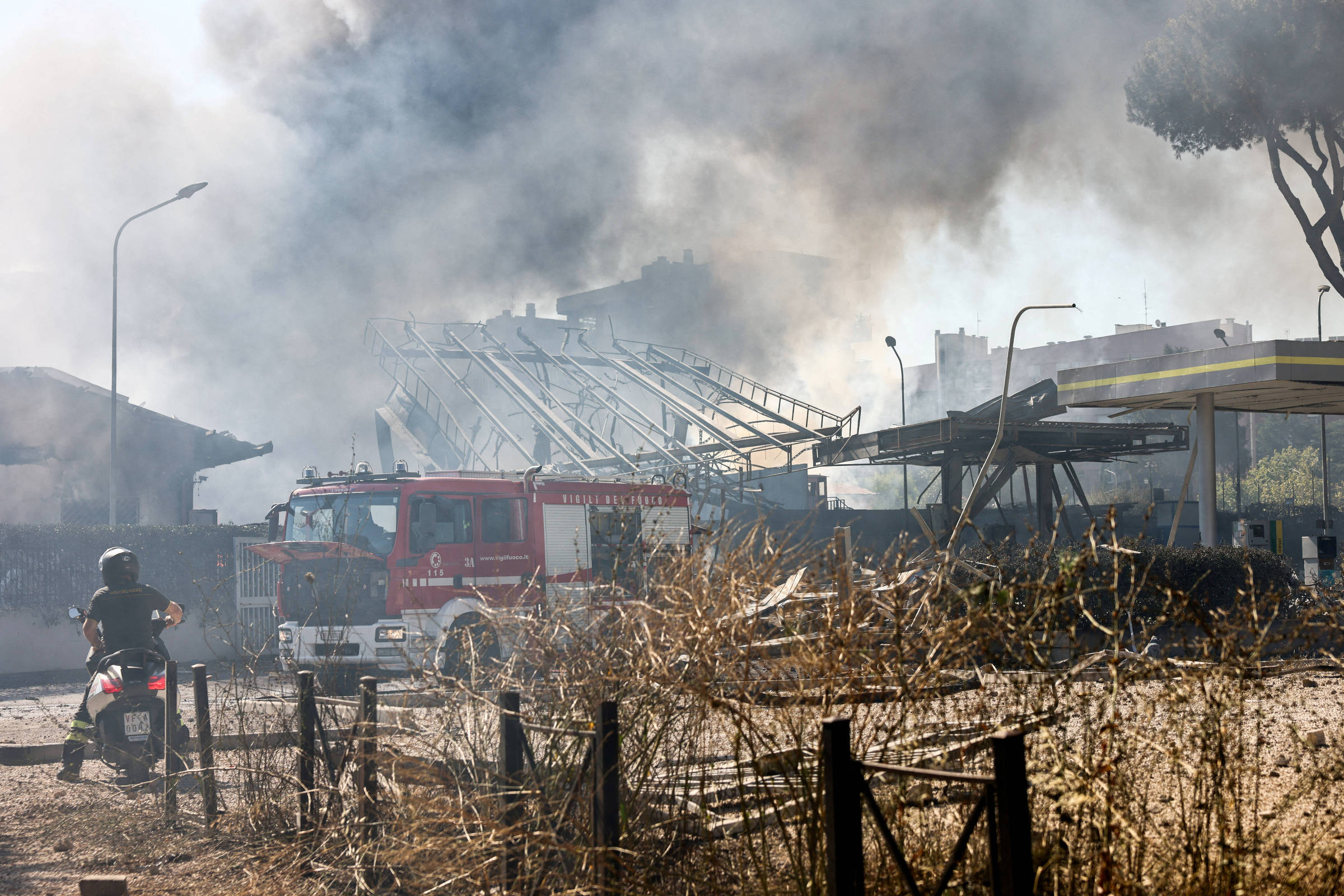 A imagem mostra um incêndio em uma área urbana, com fumaça densa subindo no ar. Um caminhão de bombeiros está presente, próximo a uma estrutura danificada. No primeiro plano, há um motociclista observando a cena. O ambiente parece seco e há vegetação morta ao redor.
