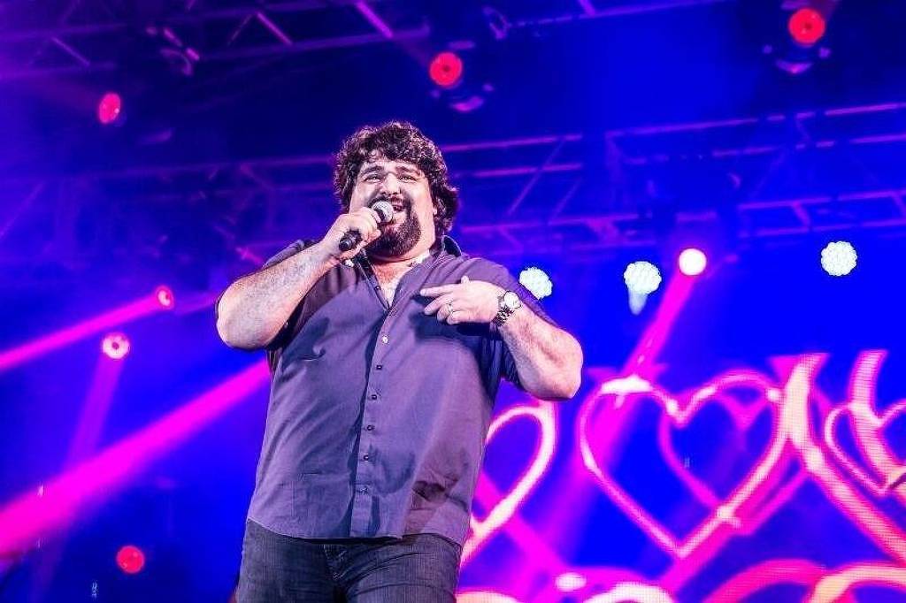 Um cantor está se apresentando em um palco iluminado com luzes coloridas. Ele segura um microfone e parece estar interagindo com o público. O fundo é decorado com luzes em forma de corações, criando um ambiente festivo. O cantor usa uma camisa escura e tem cabelo cacheado e volumoso.