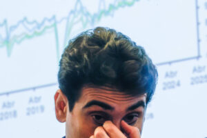 Um homem com cabelo escuro e ondulado está em primeiro plano, com uma expressão de preocupação, cobrindo parcialmente o rosto com a mão. Ao fundo, há um gráfico em uma tela, mostrando linhas de dados que variam ao longo do tempo, com datas marcadas em abril de 2018 e abril de 2020.