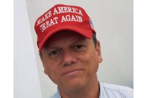 Um homem está olhando para a câmera, usando um boné vermelho com a frase