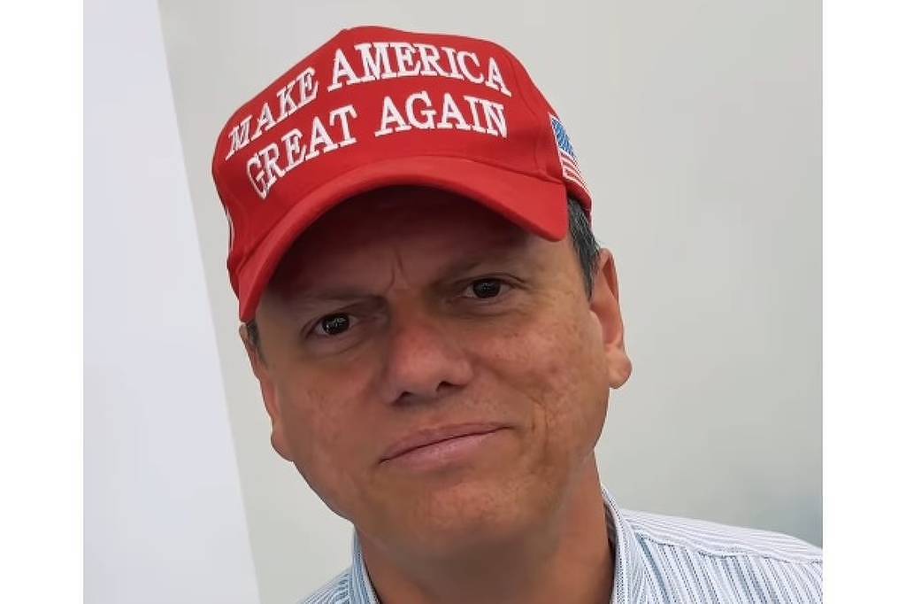 Um homem está olhando para a câmera, usando um boné vermelho com a frase