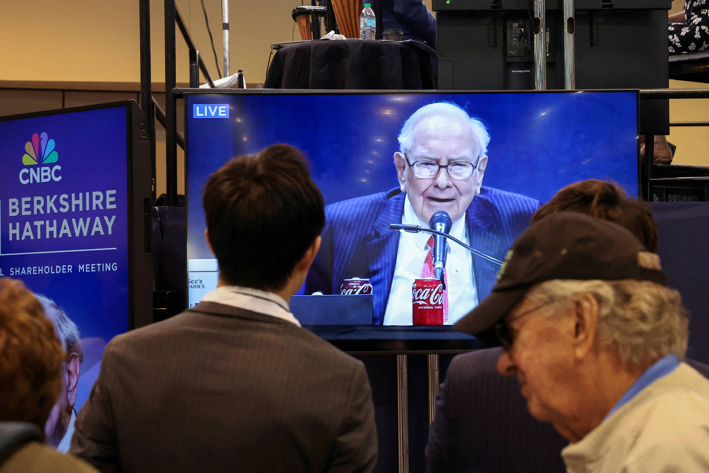 A imagem mostra um grupo de pessoas assistindo a uma transmissão ao vivo de uma reunião de acionistas da Berkshire Hathaway. Na tela, um homem idoso está falando ao microfone, com uma garrafa de Coca-Cola à sua frente. O ambiente parece ser um auditório ou sala de conferências, com banners da CNBC e da Berkshire Hathaway visíveis ao fundo.