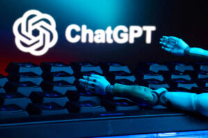 A imagem mostra um teclado de computador com algumas teclas em destaque. Em cima do teclado, há um braço robótico que parece estar se apoiando. Ao fundo, está o logotipo do ChatGPT, iluminado em uma cor clara, contrastando com um fundo azul escuro.