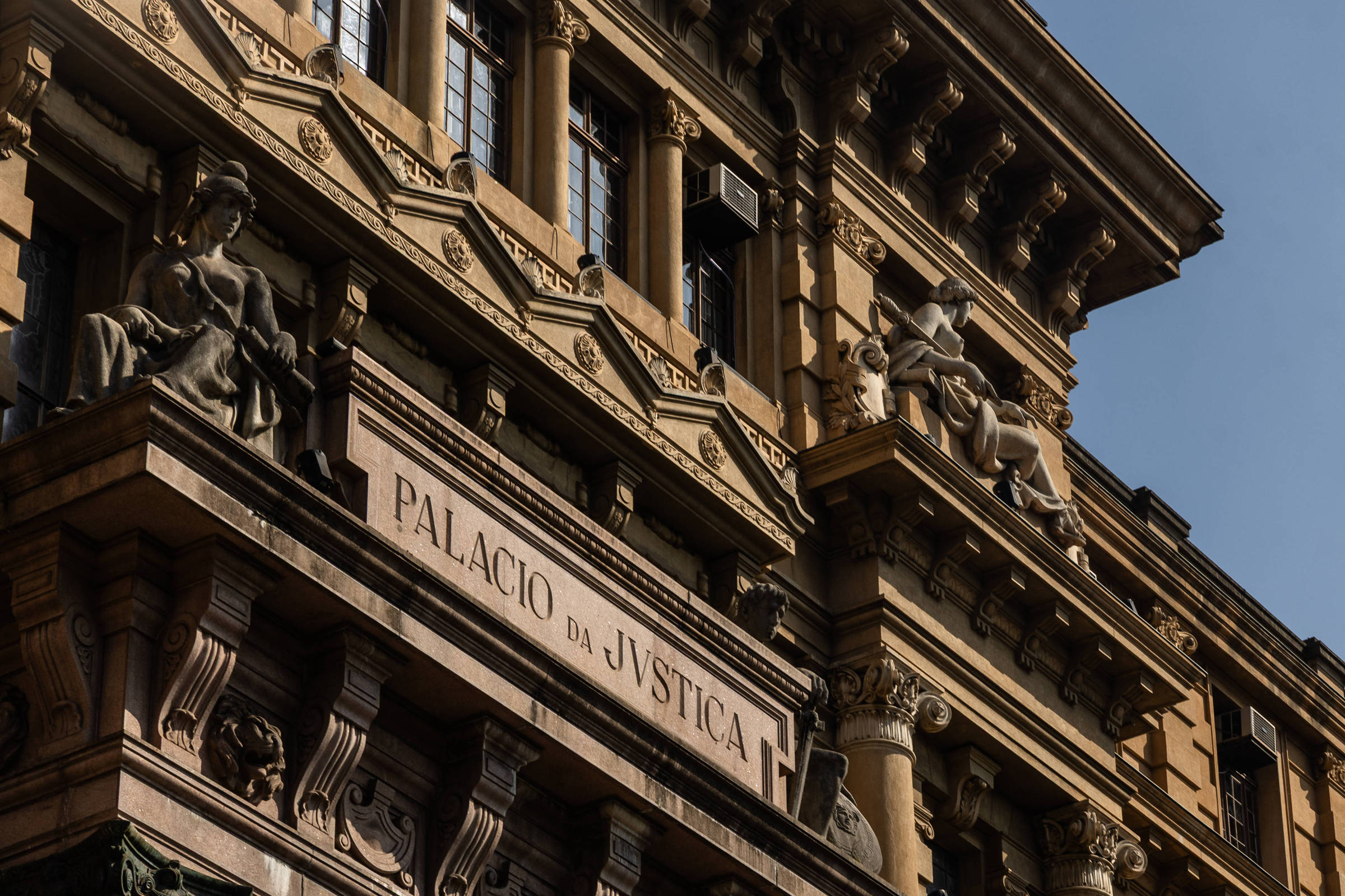 A imagem mostra a fachada do Palácio da Justiça, um edifício imponente com detalhes arquitetônicos elaborados. Na parte superior, há estátuas que representam figuras alegóricas. O nome