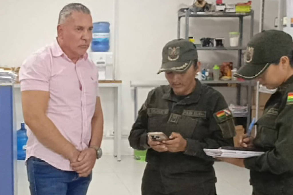 A imagem mostra um homem de camisa rosa, com expressão séria, em pé, enquanto duas mulheres em uniformes militares, uma delas usando um boné, estão ao seu redor. Uma das mulheres está olhando para um celular, enquanto a outra faz anotações em um bloco. O ambiente parece ser um escritório, com prateleiras ao fundo e alguns objetos visíveis.