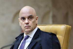 Um homem calvo, com pele clara, está sentado em uma cadeira amarela. Ele usa um terno escuro e uma gravata roxa. O fundo é desfocado, mas parece ser um ambiente formal, possivelmente um tribunal. O homem tem uma expressão séria e está olhando para frente.