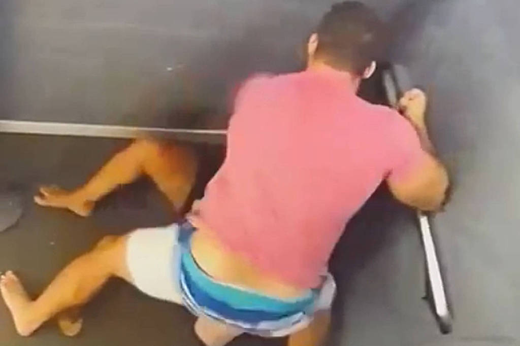 A imagem mostra duas pessoas em um espaço confinado, aparentemente lutando. Uma pessoa está em pé, vestindo uma camiseta rosa e shorts claros, enquanto a outra está no chão, com as pernas visíveis. O ambiente parece ser uma cabine ou um espaço pequeno e fechado.