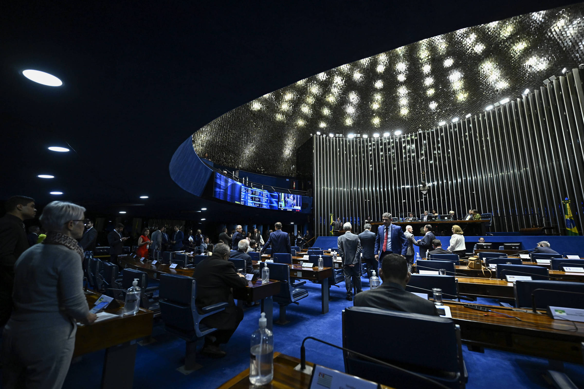 A imagem mostra o plenário do Senado Federal do Brasil, com senadores em pé e outros sentados em suas cadeiras. O ambiente é iluminado por uma iluminação moderna e o teto é curvo, com um design contemporâneo. Ao fundo, há painéis eletrônicos e a bandeira do Brasil. As mesas são de madeira e há garrafas de água sobre elas.