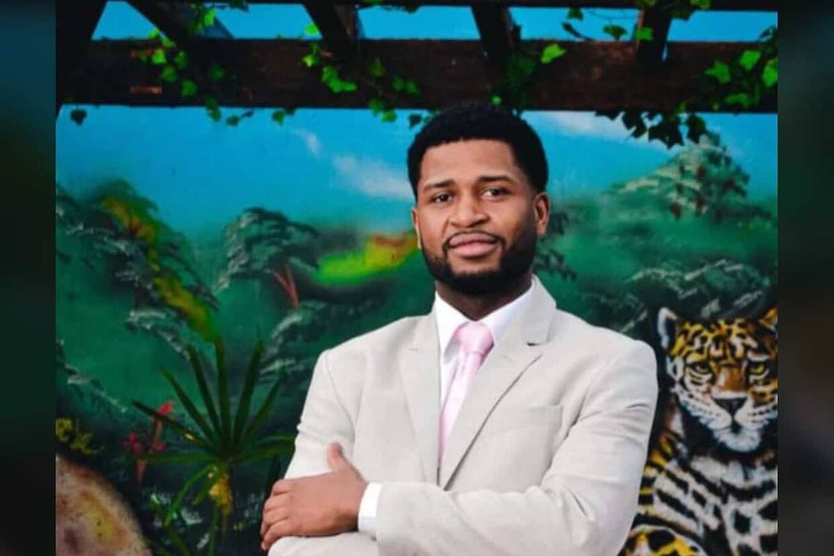 Um homem está posando em frente a um mural colorido que apresenta uma onça e vegetação tropical. Ele está vestido com um terno claro e uma gravata rosa, com os braços cruzados e um sorriso confiante. O fundo é vibrante, com tons de verde e detalhes da fauna.