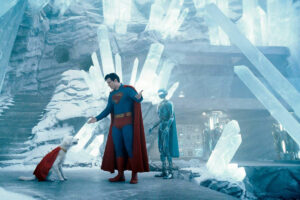 A imagem mostra um personagem vestido como super-herói, com um traje azul e uma capa vermelha, interagindo com um cachorro que também usa uma capa vermelha. Ao fundo, há uma estrutura de cristais de gelo e um robô prateado. O ambiente é gelado e iluminado, com uma atmosfera futurista.