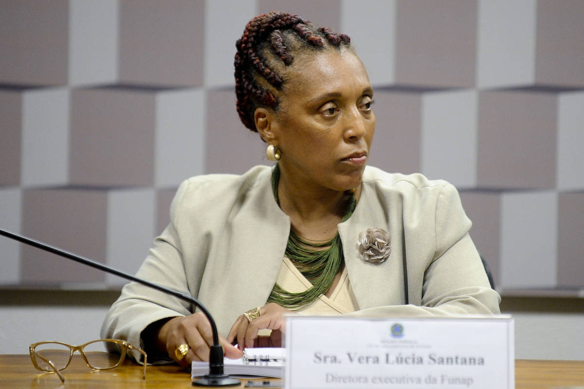 A imagem mostra uma mulher sentada à mesa em um ambiente formal. Ela tem cabelo trançado e usa um blazer claro. Na mesa, há um nome em uma placa que diz