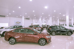 A imagem mostra um showroom de carros com várias opções de veículos expostos. Em primeiro plano, há um carro vermelho, enquanto ao fundo são visíveis outros carros, incluindo um SUV preto e um carro branco. O ambiente é bem iluminado, com piso brilhante e paredes brancas, criando uma atmosfera moderna e limpa.