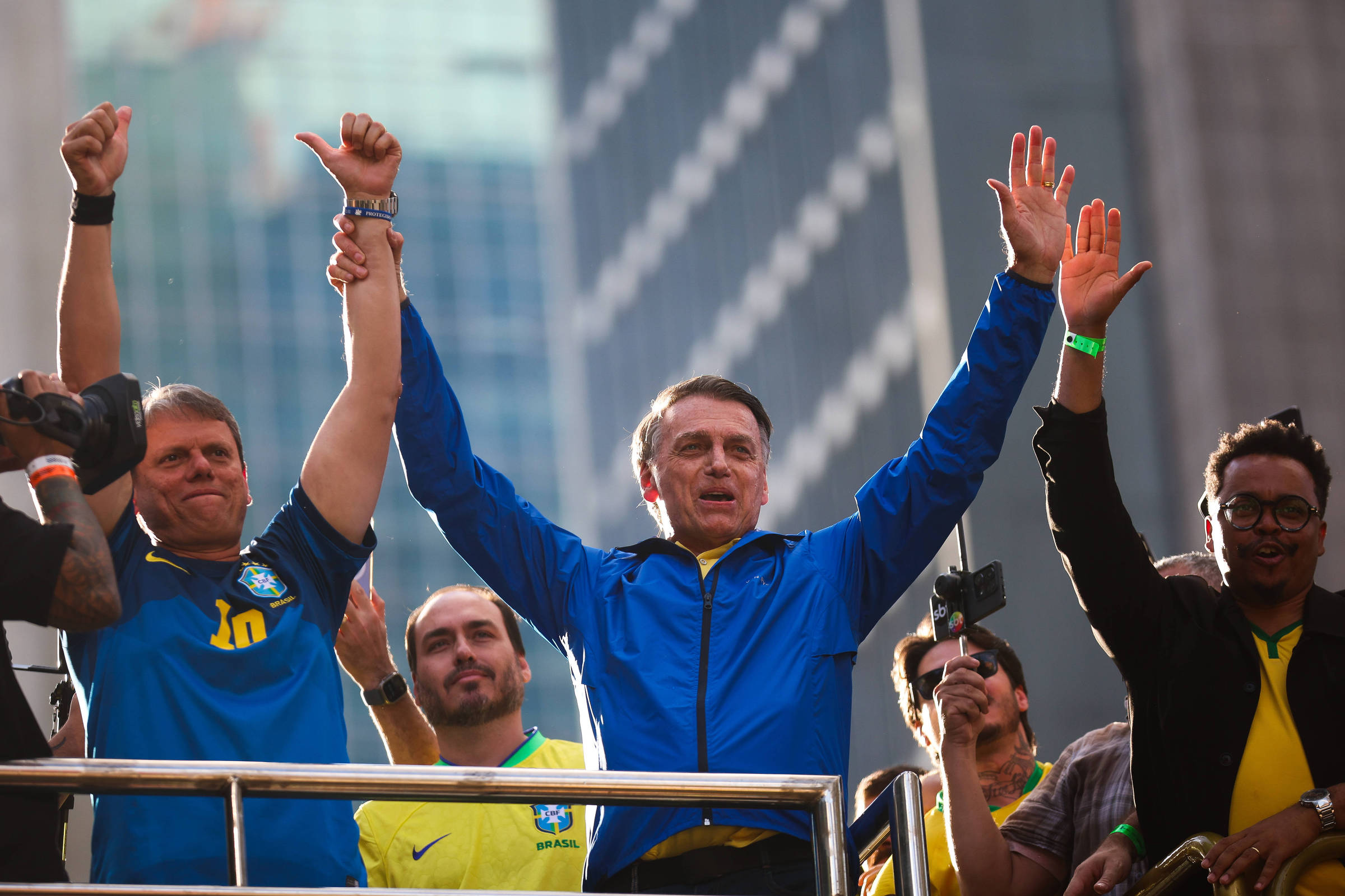 Um grupo de homens está em um palco, com um deles levantando as mãos em sinal de vitória. Eles estão vestidos com camisetas da seleção brasileira de futebol. O homem no centro, que usa uma jaqueta azul, parece estar se dirigindo a uma multidão. Ao fundo, há edifícios altos e uma atmosfera de celebração.