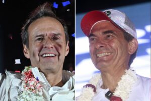 A imagem mostra dois homens sorrindo, um ao lado do outro. O homem à esquerda tem cabelo escuro e usa uma camisa clara, adornada com colares de flores. O homem à direita usa um boné vermelho e uma camisa branca, também com colares de flores. Ambos parecem estar em um evento festivo, com confetes coloridos ao fundo.